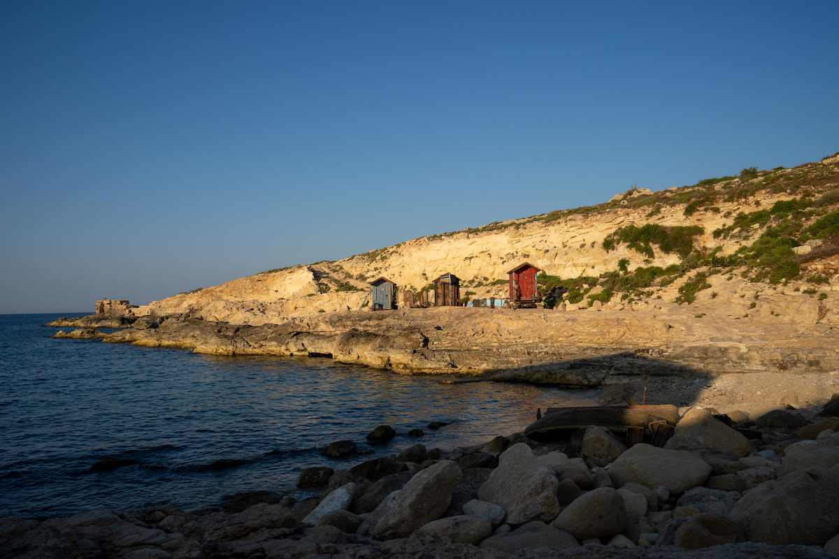 Small wooden huts on a rocky coastline with clear blue sea and sunlit cliffs, under a clear sky, with some greenery scattered on the cliffs.