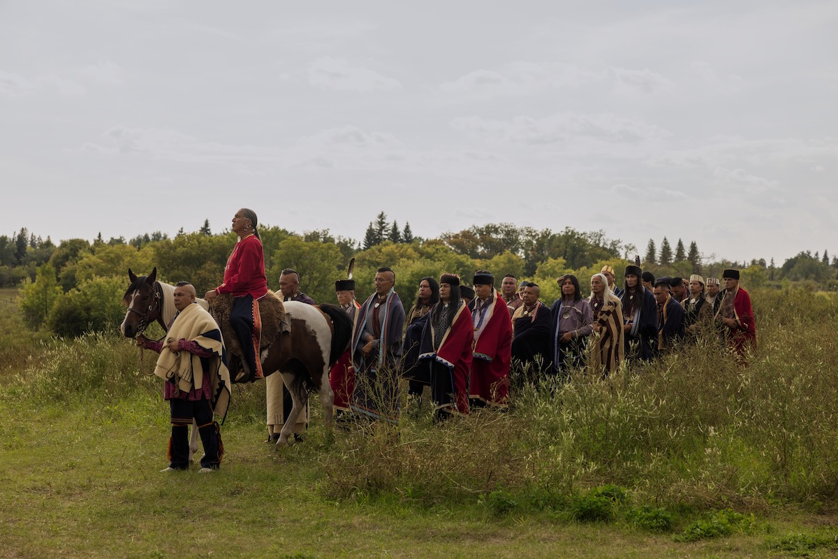 Group of people in historical clothing walking through grassy field, one person on horseback, surrounded by trees and overcast sky in a natural outdoor setting.