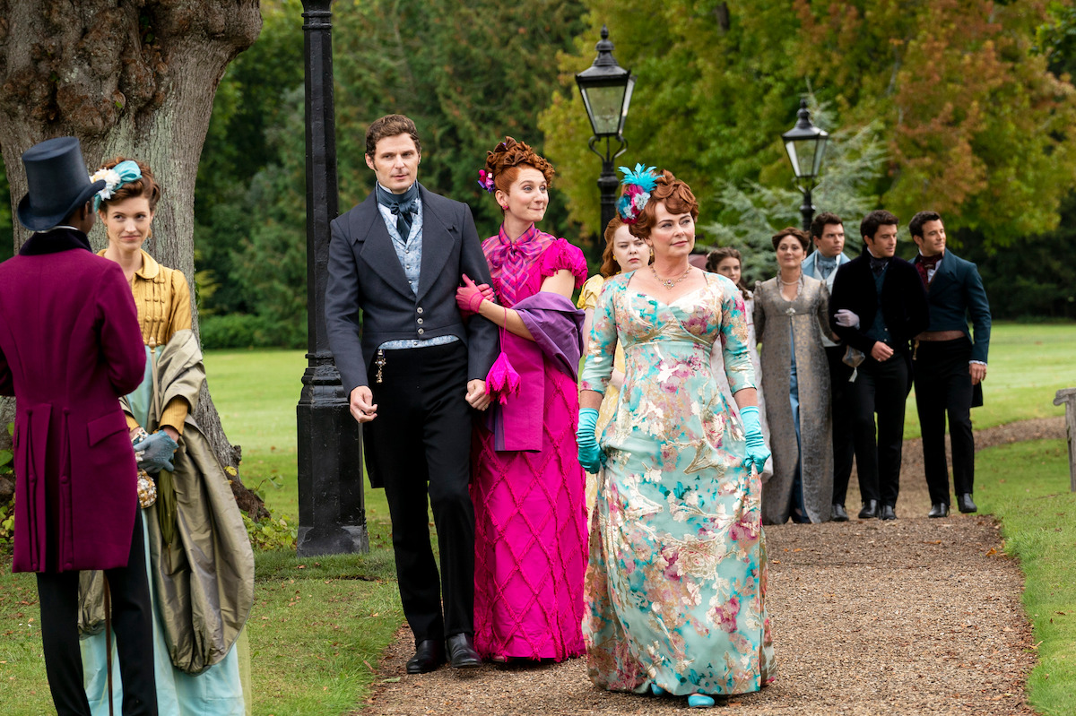People in 19th-century period costumes walk and talk on a gravel path in a lush garden with trees and old-fashioned street lamps, some looking formal and elegant, suggesting a social or historical event outdoors.