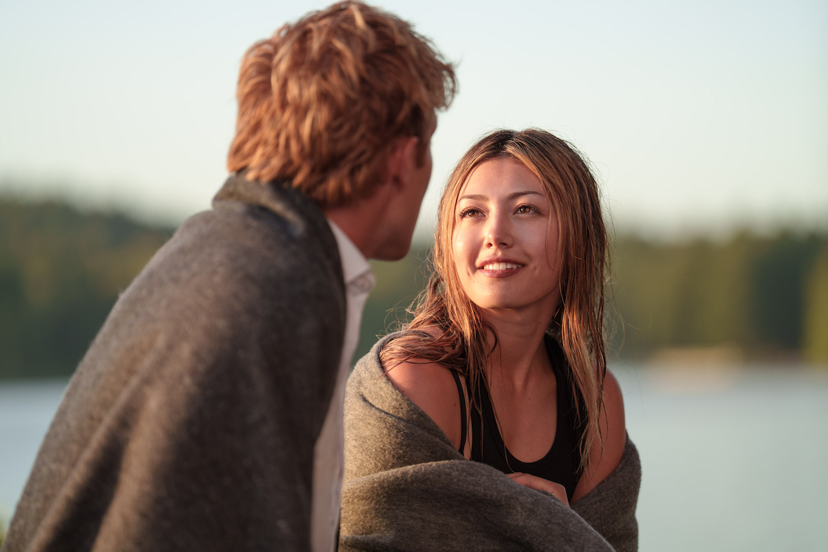 Two people wrapped in blankets sit closely together outdoors by a lake, surrounded by trees, sharing a warm moment in the soft evening light.