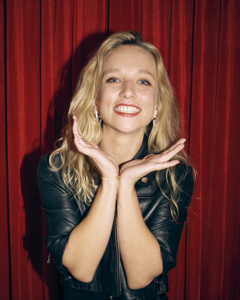 Smiling woman with long blonde hair in a black leather jacket posing with hands framed under her chin, standing in front of a red curtain background, indoor setting with bright lighting.