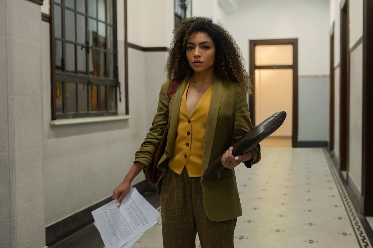 A atriz Any Gabrielly, de cabelos cacheados e roupas formais, aparece em um corredor segurando uma pasta com documentos