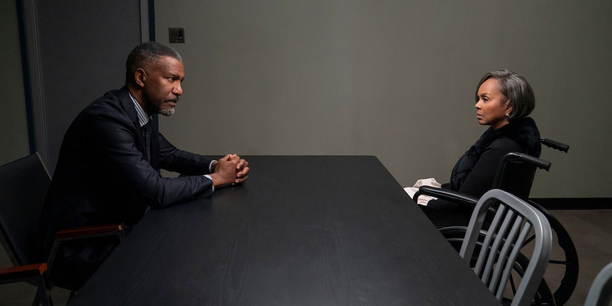 A man and a woman sit across from each other at a black table in a sparse, dimly lit room; the woman is in a wheelchair. The setting suggests a serious conversation, possibly an interrogation or meeting.