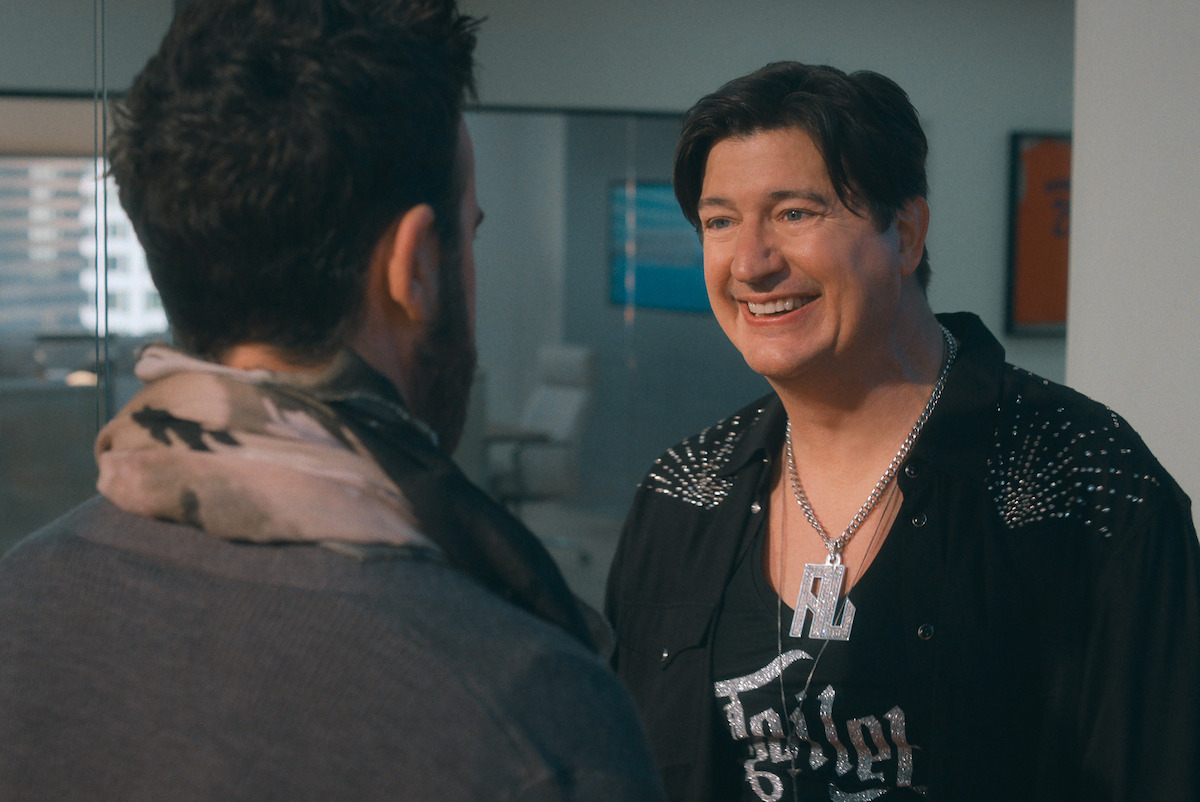 Two men having a conversation in a modern office, one facing the camera and smiling, the other seen from behind, with glass walls and office furniture in the background.