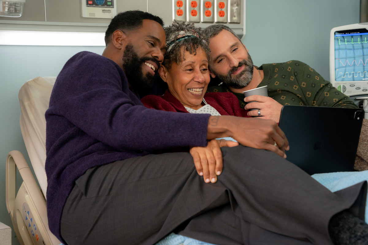 Three people sit together smiling on a hospital bed, enjoying time together. Medical equipment is visible in the background, suggesting a supportive and caring hospital environment.