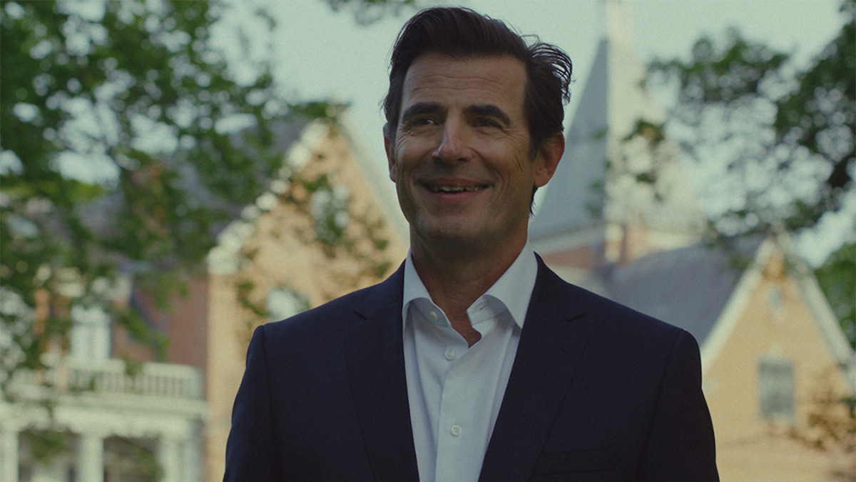 A smiling man in a dark suit stands outdoors in front of a large brick building with trees and a steeple visible in the background, suggesting a formal or academic setting on a sunny day.