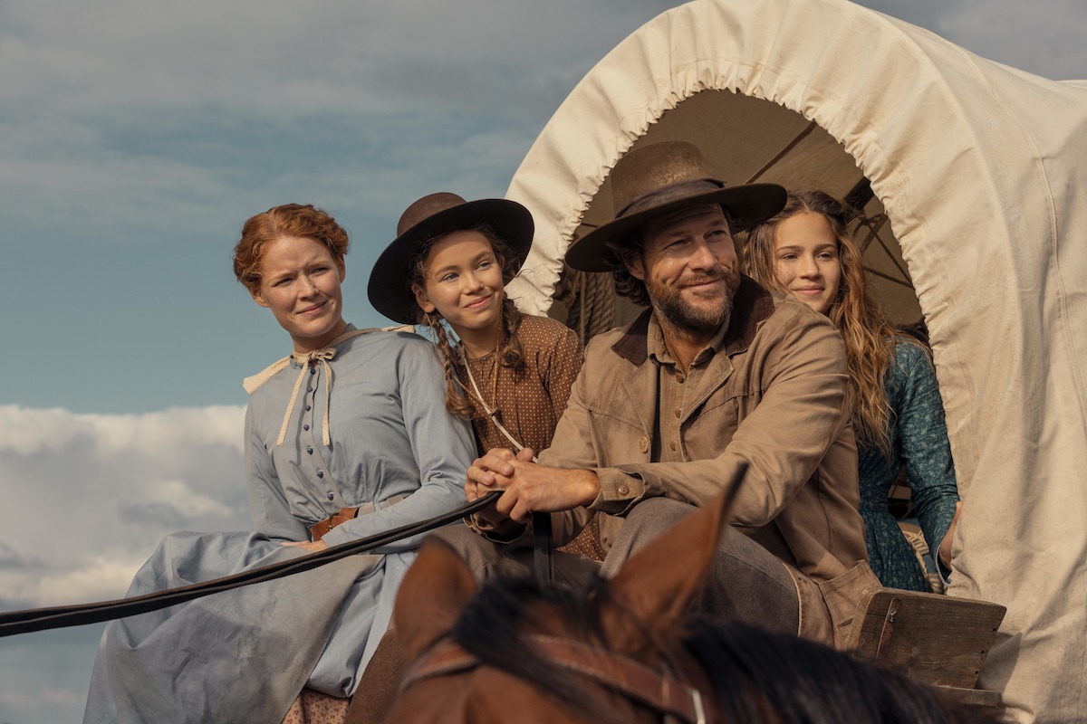 A man and three women in pioneer-era clothing ride a horse-drawn covered wagon outdoors, under a blue sky with clouds, suggesting a journey or travel in a historical rural setting.