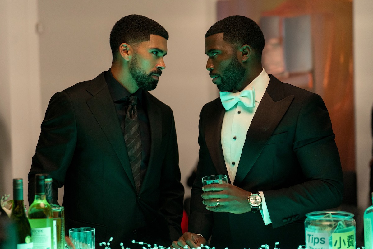 Two men in formal black suits stand at a bar, engaged in a serious conversation. The setting appears to be an upscale event or party, with bottles, drinks, and ambient lighting on the bar counter.