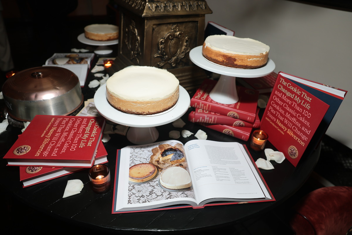 Cheesecakes on stands and an open cookbook arranged on a round table with red books, candles, and white rose petals, creating a cozy and inviting dessert display in an intimate indoor setting.