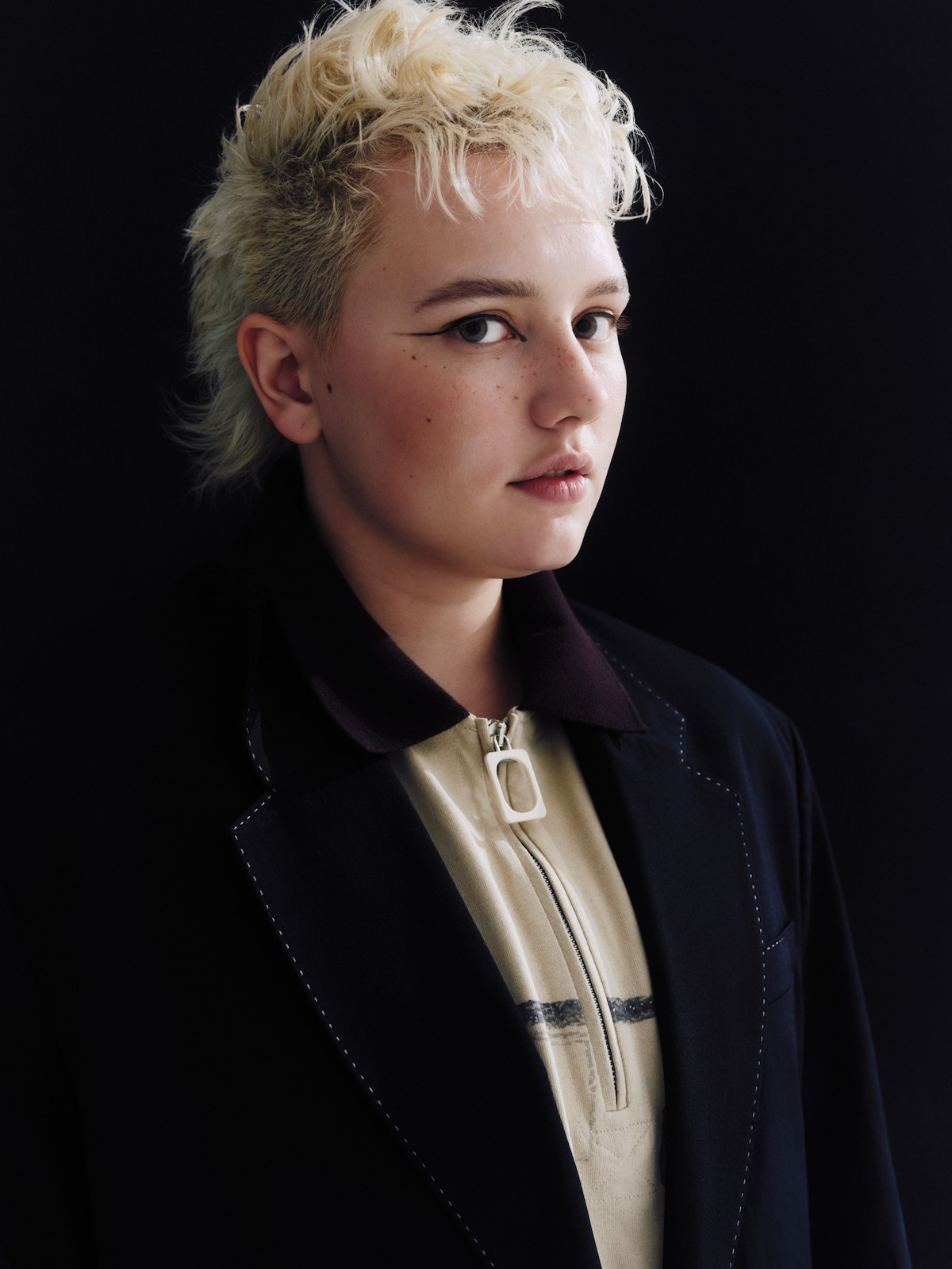 Person with short, bleached blond hair and a mullet wearing a dark jacket and beige zip-up shirt, standing against a plain dark background with soft, even lighting.