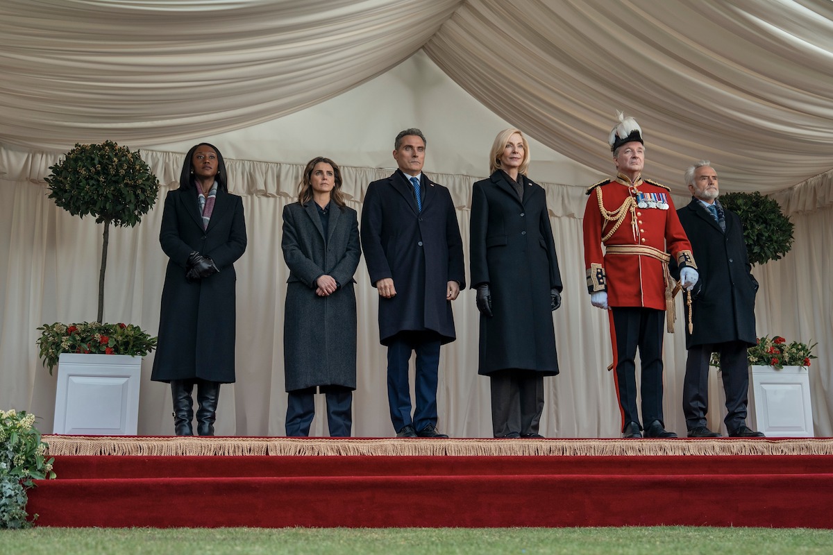 Six formally dressed people stand on a red-carpeted stage under a draped white canopy, flanked by potted plants; one wears a ceremonial military uniform, the others wear dark coats, appearing to attend an official or formal outdoor event.