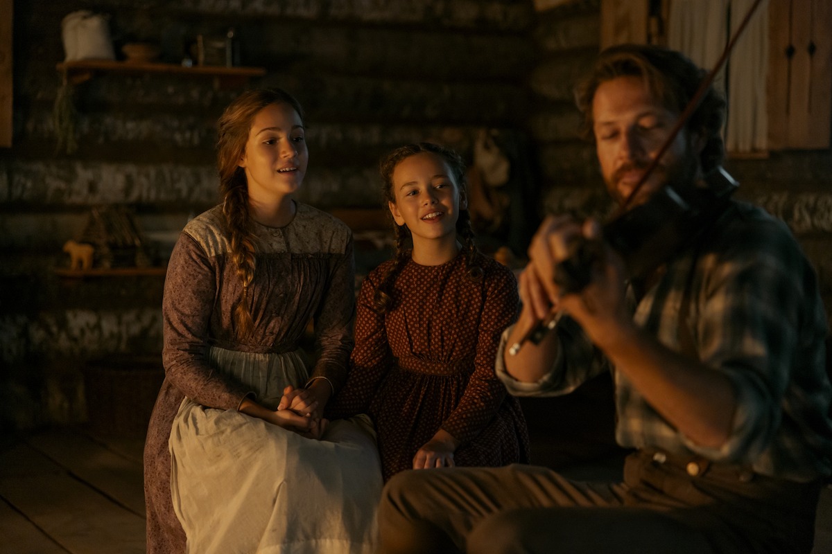 A man plays the violin for a woman and a young girl inside a rustic wooden cabin, creating a warm, cozy atmosphere with vintage clothing and soft lighting.