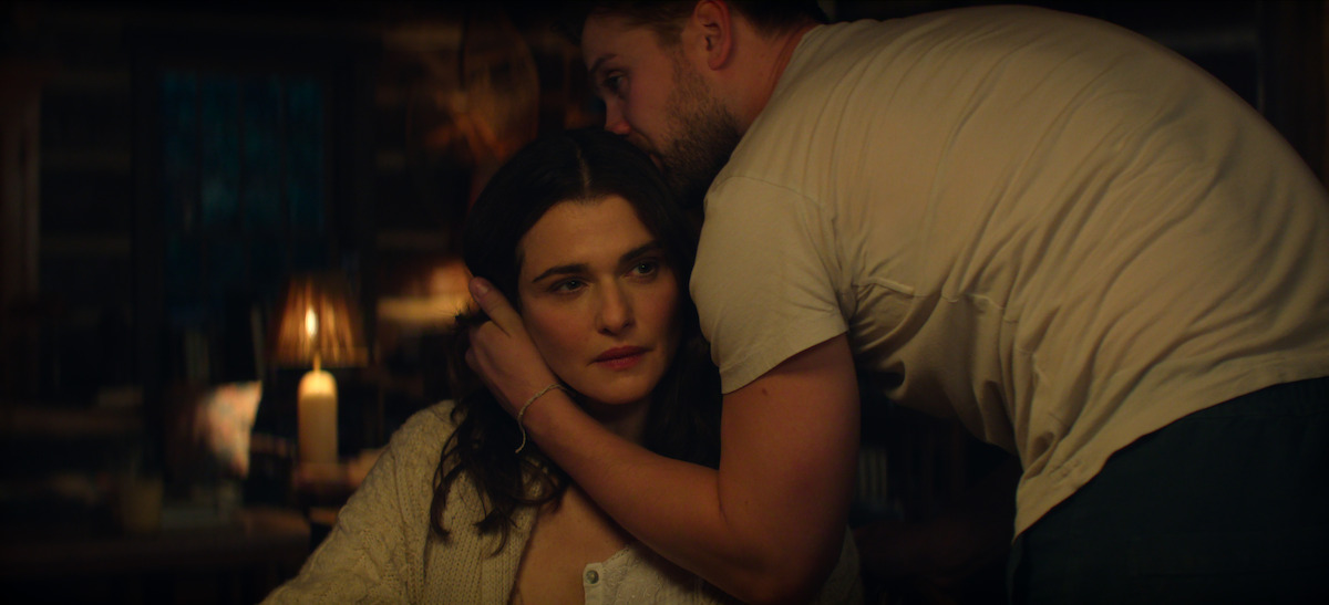 Rachel Weisz as The Protagonist sitting at a table in a softly lit, cozy room with a lamp in the background; Leo Woodall as Vladimir leans in closely, gently touching her head and whispering, creating an intimate, contemplative atmosphere.