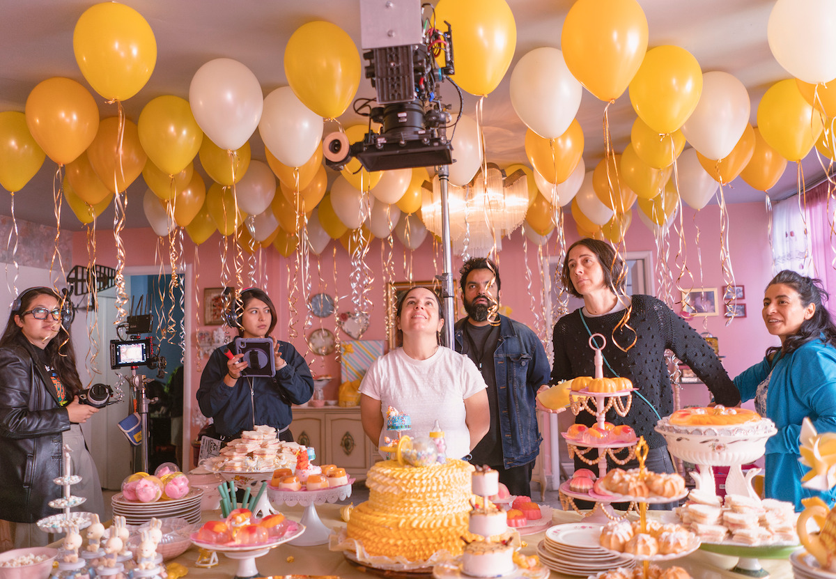A group of people filming at a festive indoor party scene with yellow and white balloons, cakes, and desserts on a decorated table, surrounded by film equipment and cameras under warm lighting.