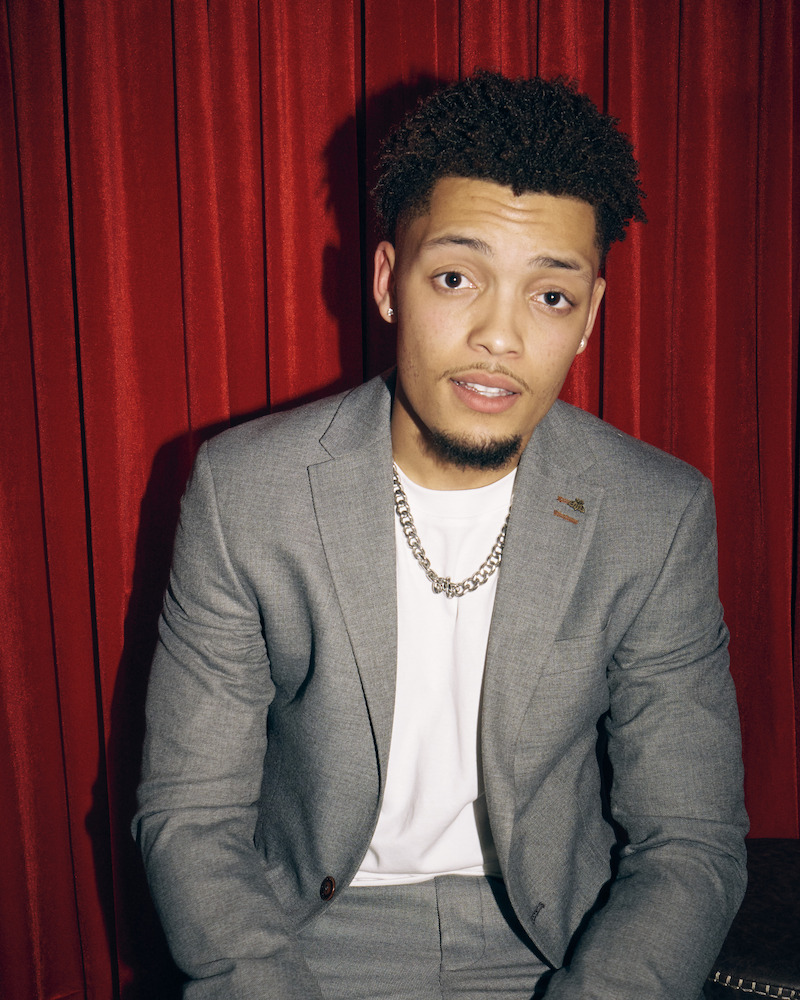 Man in a light grey suit and white shirt wearing a chain necklace, sitting in front of red curtains, looking at the camera in a formal indoor setting.