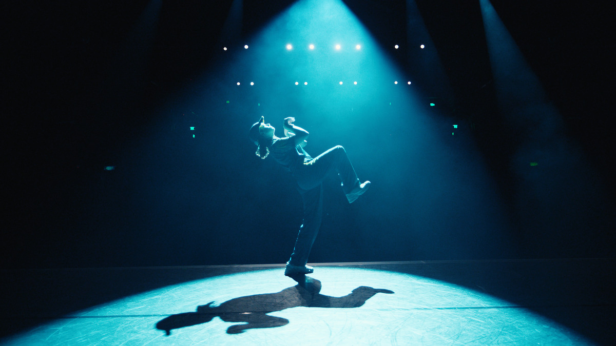 A person dancing alone on stage under dramatic blue spotlight, surrounded by darkness with stage lights overhead, casting a strong shadow on the floor in a theatrical performance setting.