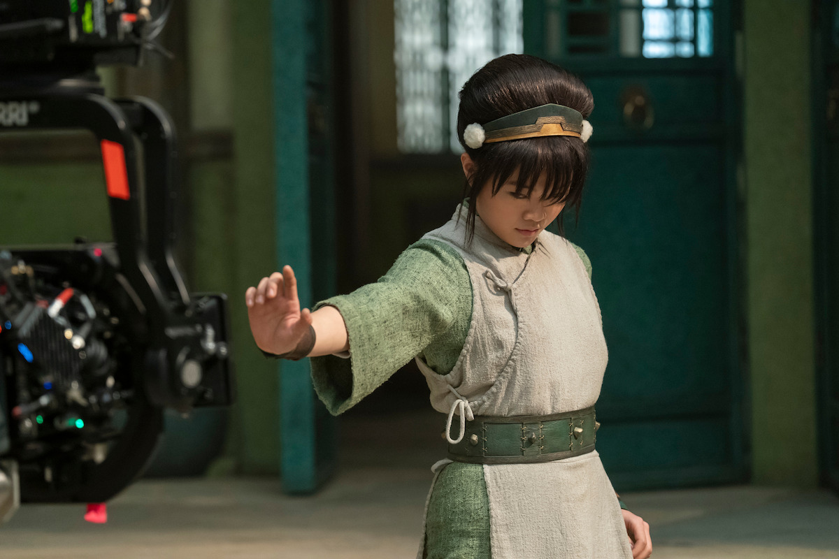 Miyako as Toph in a green and gray costume poses with one arm extended in front of a film set, with a camera rig visible. The environment features teal doors and green walls, suggesting a fantasy or historical production setting.