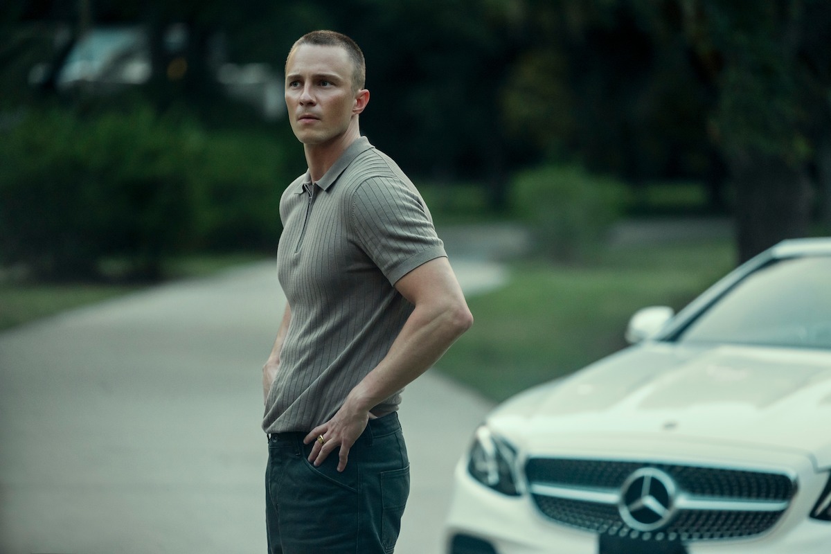 A man stands outdoors on a driveway next to a white Mercedes-Benz car, surrounded by greenery on a cloudy day.