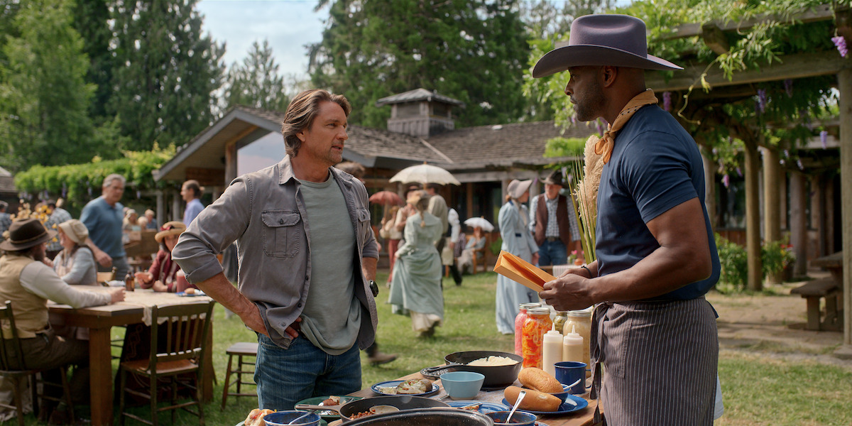 Two men talk by a food table at an outdoor gathering, one wearing a hat and apron. People in period costumes sit and walk in the background near rustic buildings, with trees and greenery surrounding the scene.