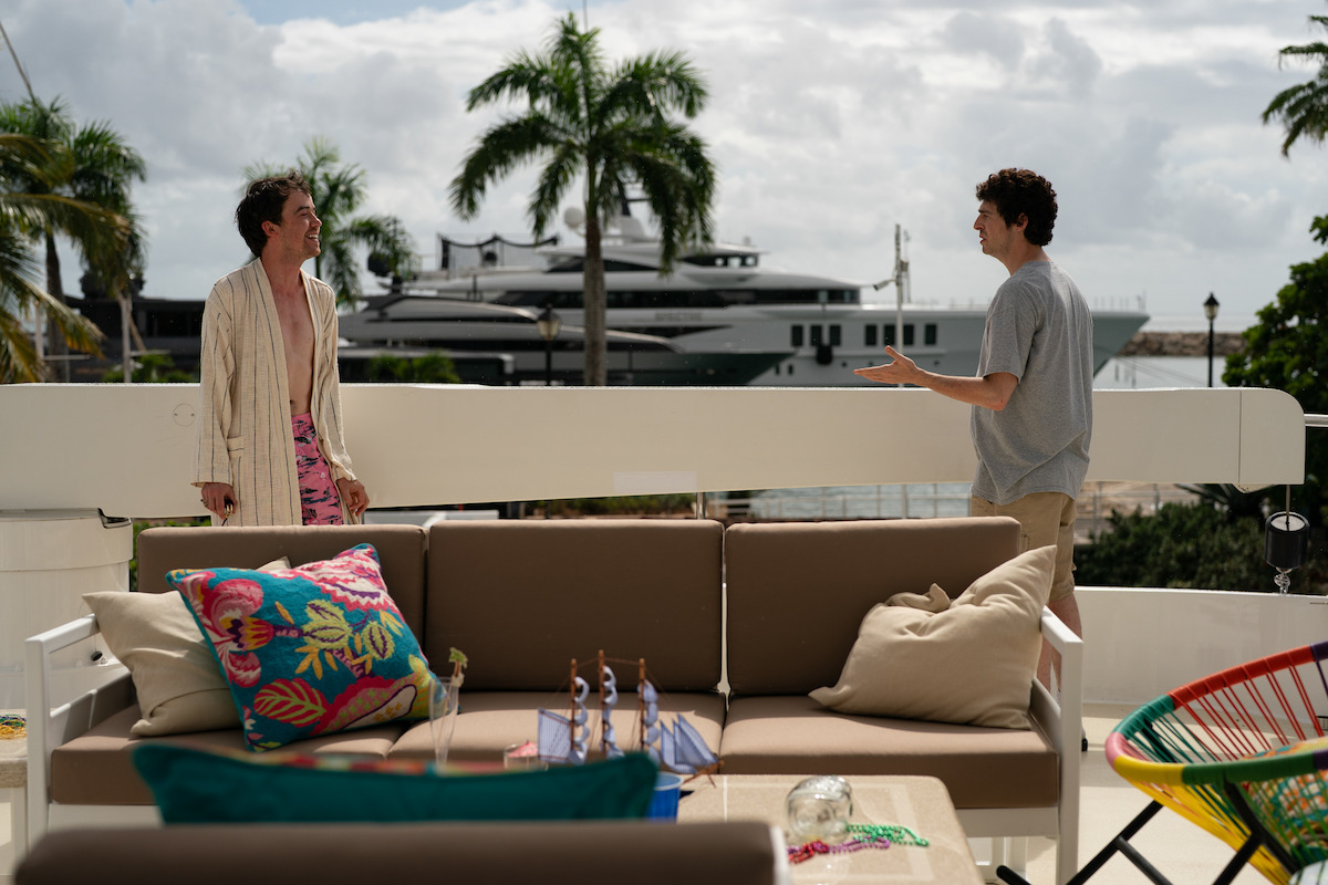 Two people are talking on a sunny outdoor patio with a sofa, colorful pillows, and a table; palm trees and a large yacht are visible in the background, creating a relaxed, tropical setting.