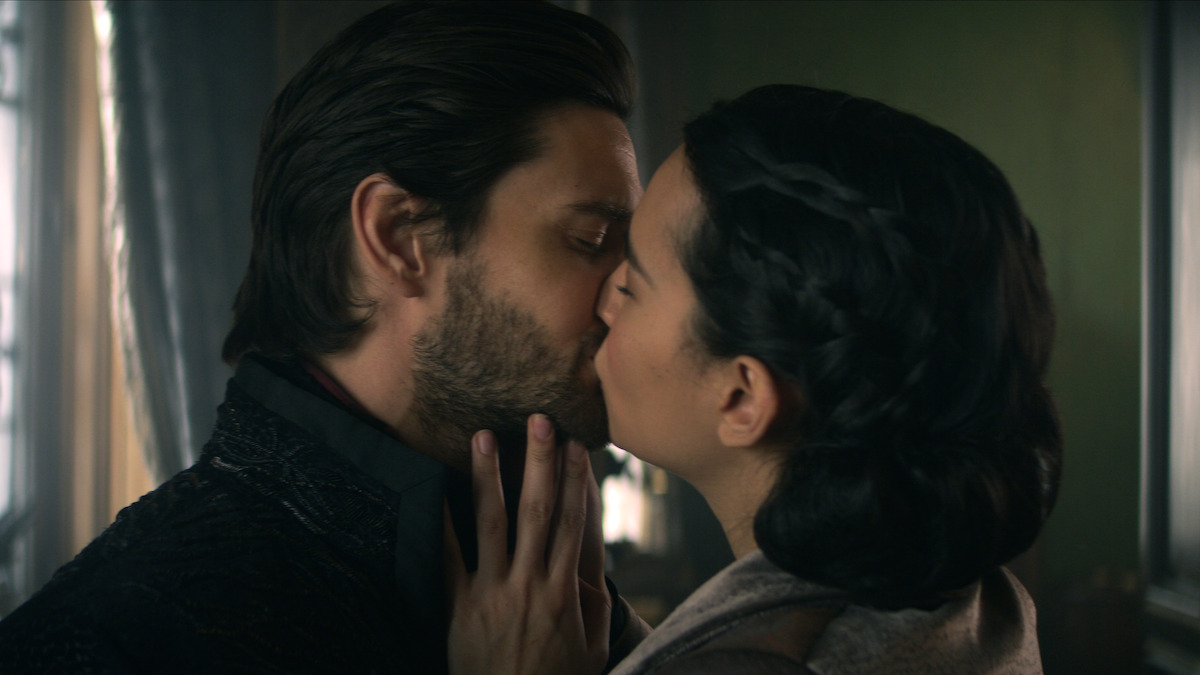 Two people share a close kiss in a softly lit, indoor setting with dark curtains and a green wall in the background. The moment is intimate and romantic, with gentle light highlighting their faces.