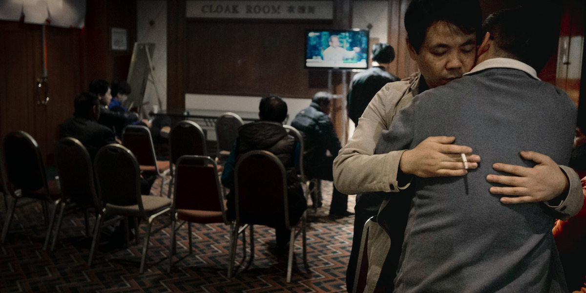 In a dark room filled with hotel chairs, two Asian men embrace each other as a television plays a news program behind them.