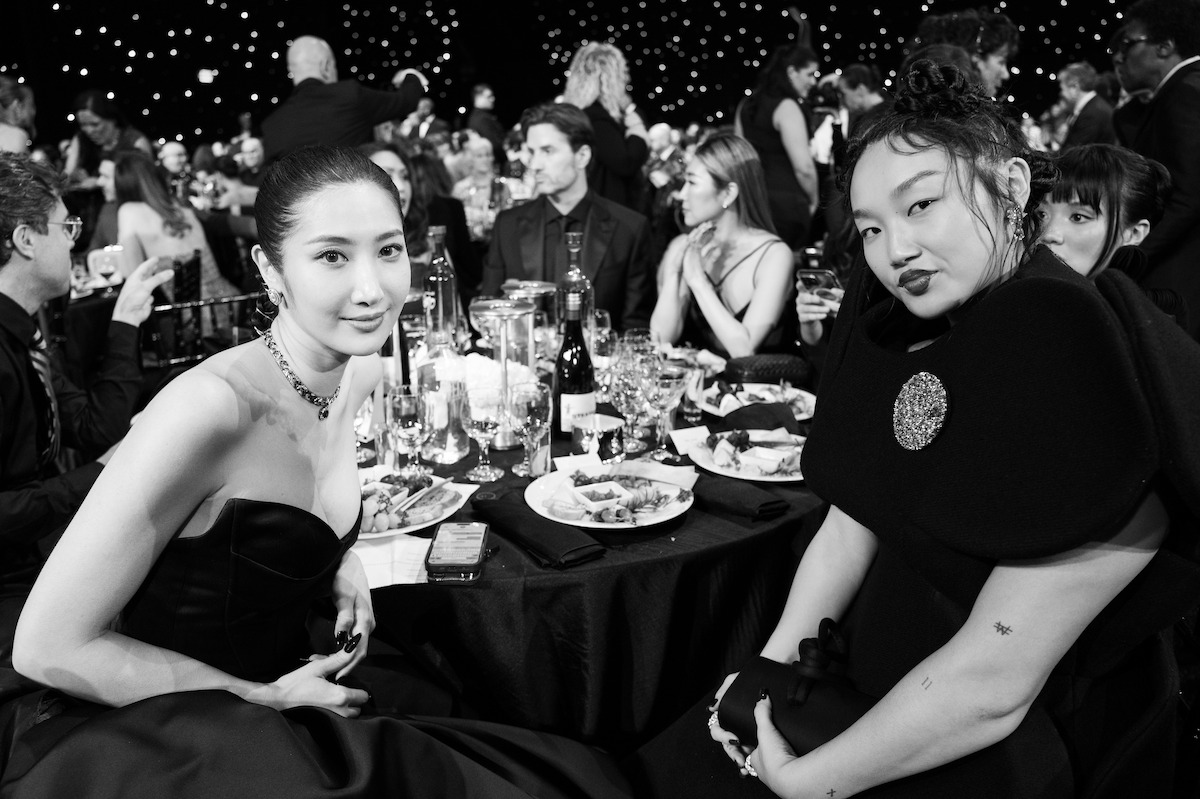 Two women dressed formally sit at a dinner table with plates, glasses, and bottles, surrounded by other guests at a glamorous event with twinkling lights in the background. Black and white photo.