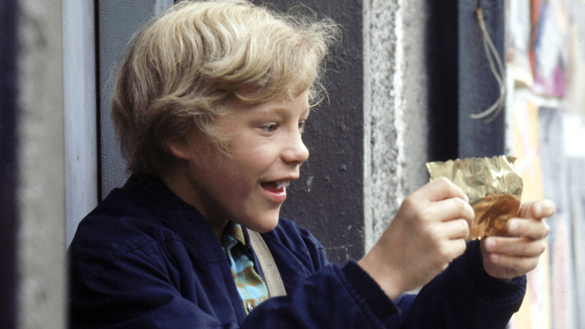 A smiling blond boy outdoors excitedly looks at a shiny golden wrapper he is holding, with a gray concrete wall and posters in the background, wearing a blue jacket.