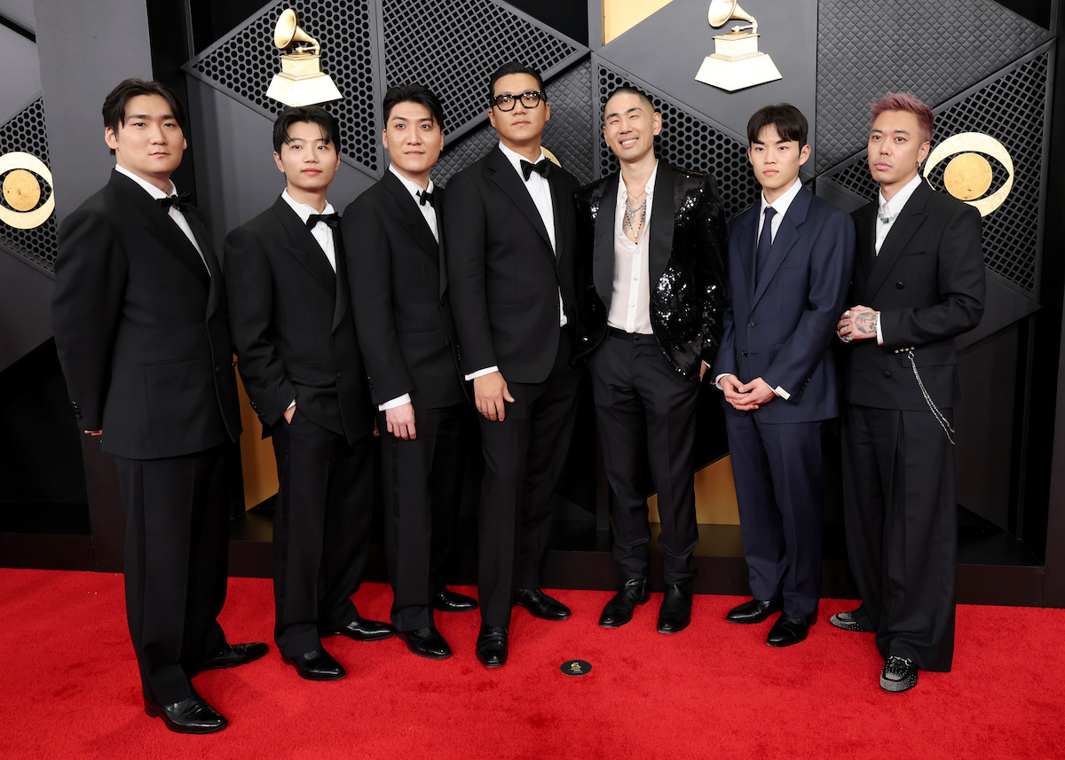 Seven men in formal suits pose together on a red carpet at an awards event, with a backdrop featuring Grammy symbols and geometric patterns behind them.