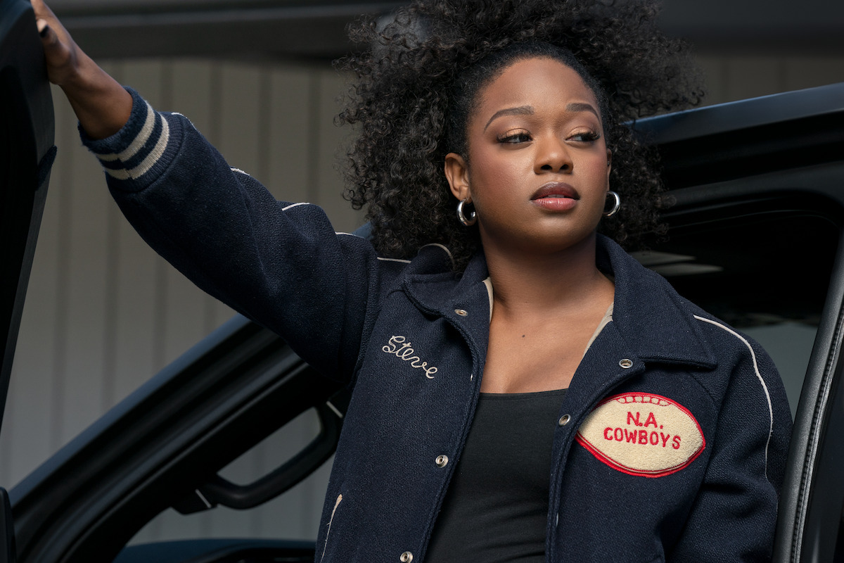 A woman with curly hair wearing a navy varsity jacket stands beside an open car door, outdoors, looking confidently to the side. The patch on her jacket reads "N.A. Cowboys" and another part says "Steve."