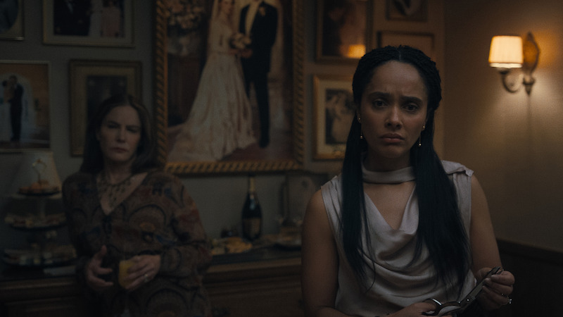 Two women stand in a dimly lit room decorated with framed photos and a wedding portrait, one holding scissors and the other with a drink, creating a tense atmosphere at what appears to be a gathering or event.