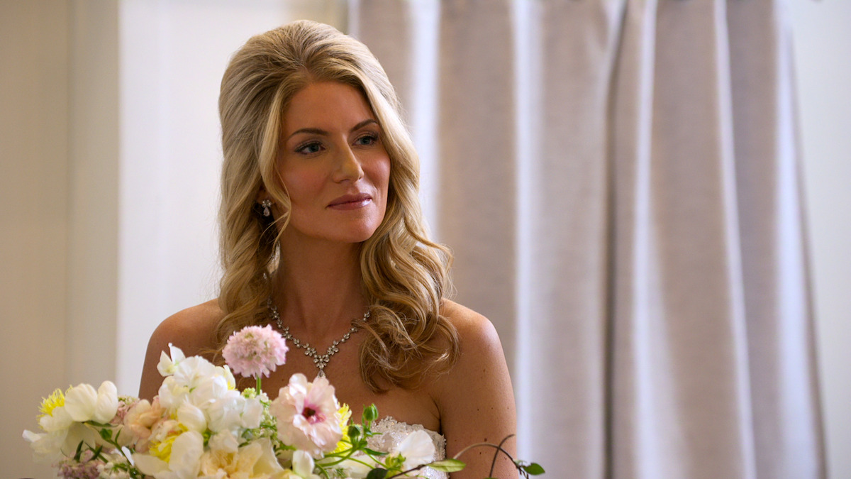 A woman in a white dress holds a bouquet of flowers indoors, standing in front of light-colored curtains, with soft lighting creating a calm atmosphere.