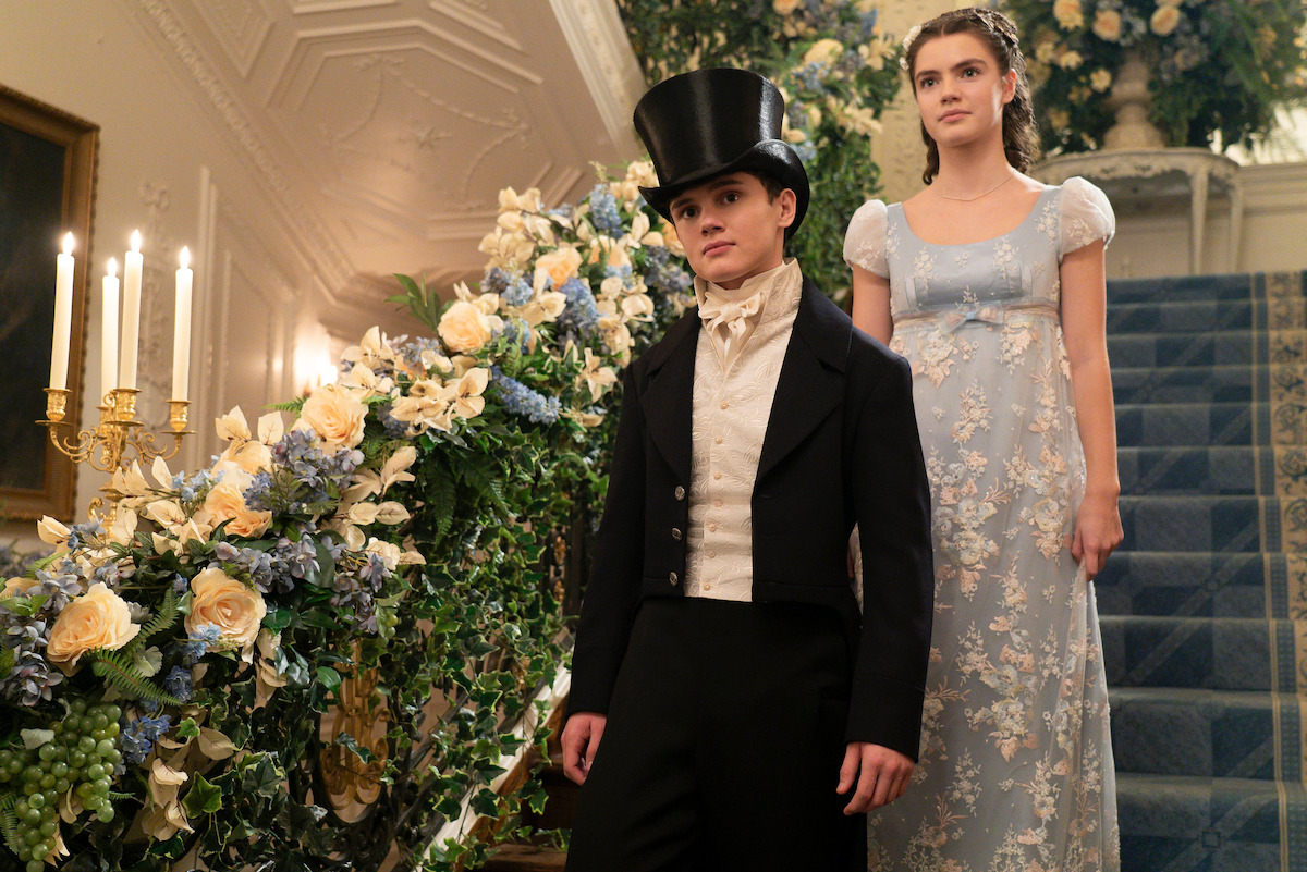 Two people in Regency-era formal attire stand on a grand staircase decorated with flowers and candles, inside an elegant setting with soft lighting.
