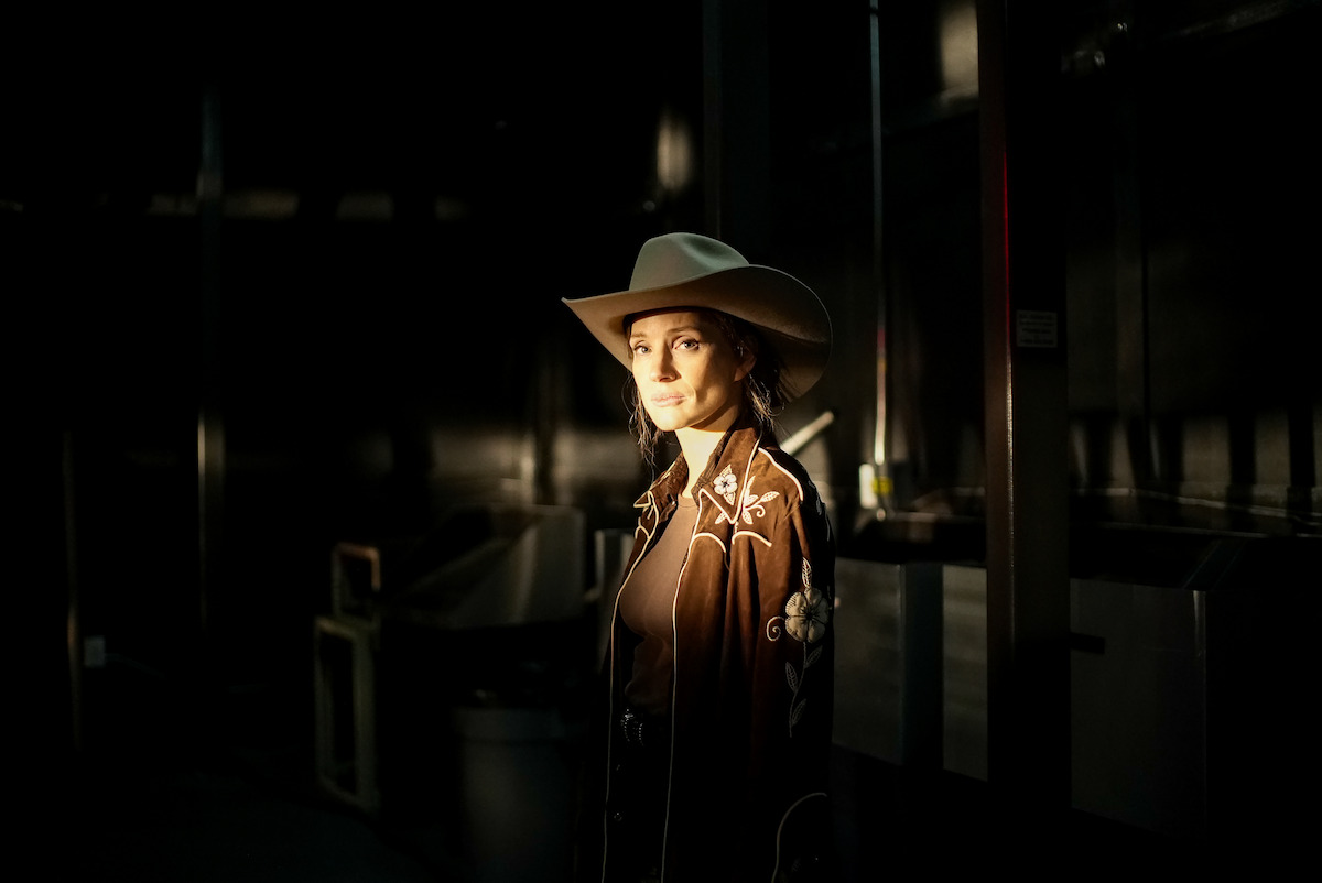 Woman in a cowboy hat and brown embroidered western shirt stands in dramatic lighting inside a dark, industrial setting with metallic walls and scattered equipment.