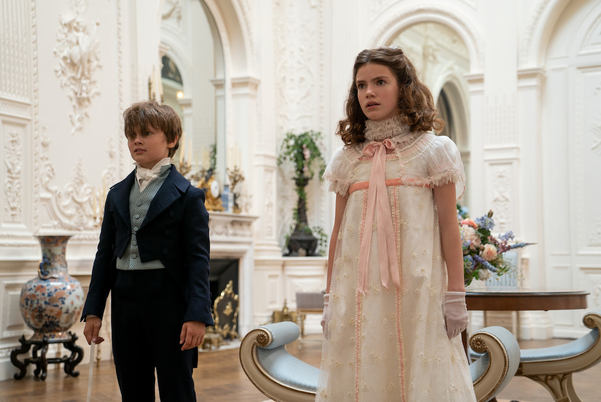 A young boy and girl in elegant period clothing stand in an ornate, white Victorian-style room with classical decor, flowers, and antique furniture.