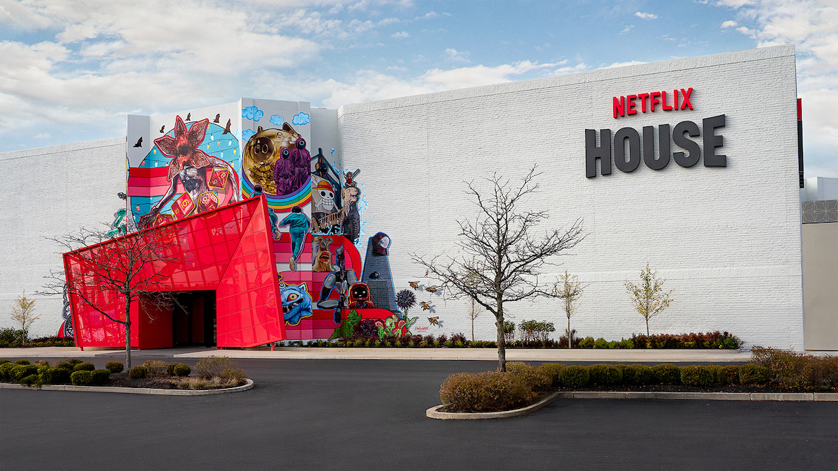 A modern white building labeled Netflix House, featuring a vibrant mural of characters, a red geometric entrance, leafless trees, landscaped bushes, and a clear sky; the mood is lively yet calm.