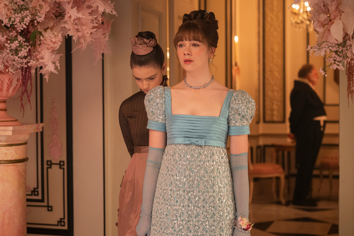 Two women in elegant period costumes stand in an ornate room with floral arrangements and chandeliers, one in a light blue gown and gloves, the other in dark attire, with a man blurred in the background.