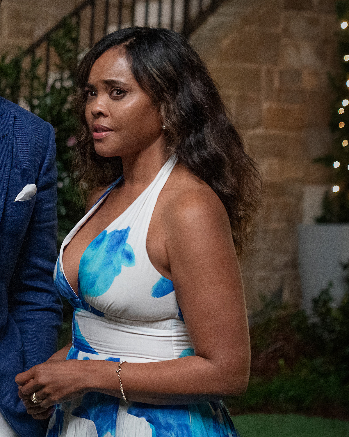 Woman in a blue and white dress standing outdoors at night, with stone walls, greenery, and string lights in the background, looking to the side with a serious expression.