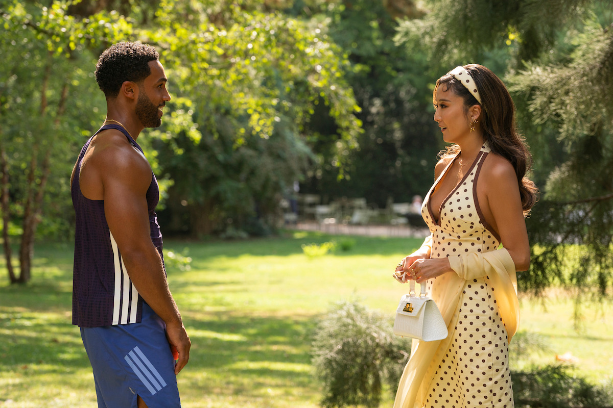 A man in athletic wear and a woman in a yellow polka dot dress stand facing each other in a sunlit park surrounded by trees, engaged in conversation on a summer day.