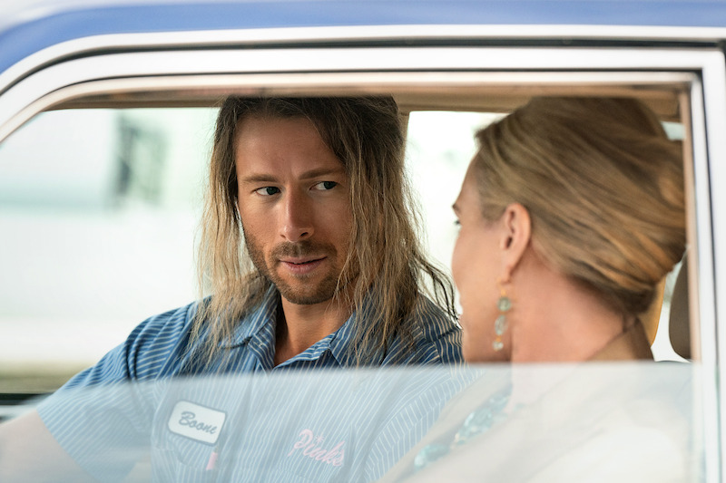 Gary Johnson (Glen Powell) undercover with a client. Johnson wears a denim top and long scraggly wig. His client is blond and has a ponytail. 