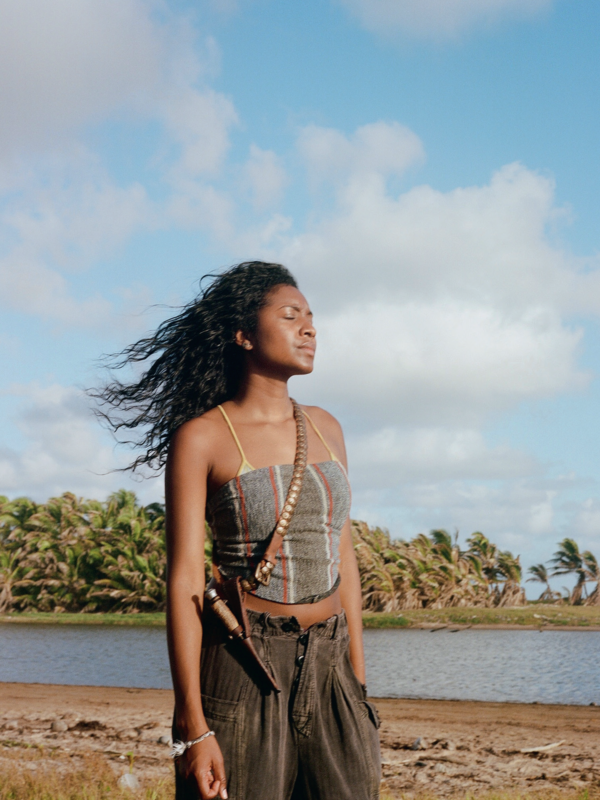 A woman stands outdoors near water and palm trees, eyes closed, enjoying the breeze under a blue sky with clouds. She wears a sleeveless top and dark pants, with a strap over her shoulder. Natural, serene tropical landscape.
