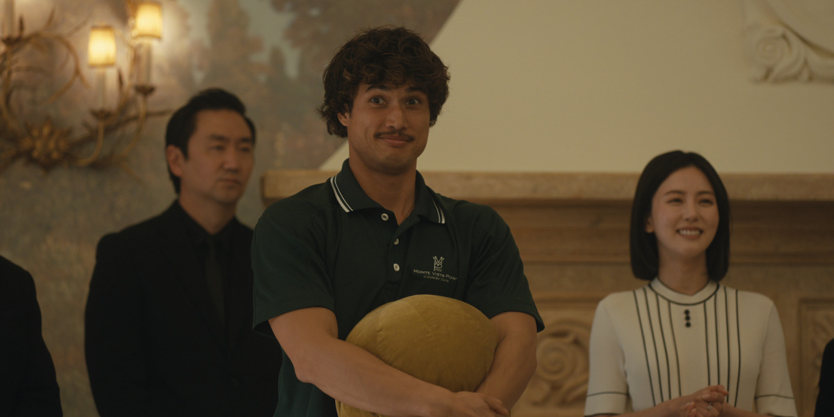 Three people standing indoors; a man in a green polo shirt holding a round yellow pillow, flanked by a man in black and a smiling woman in a white dress; ornate wall and lighting in the background.