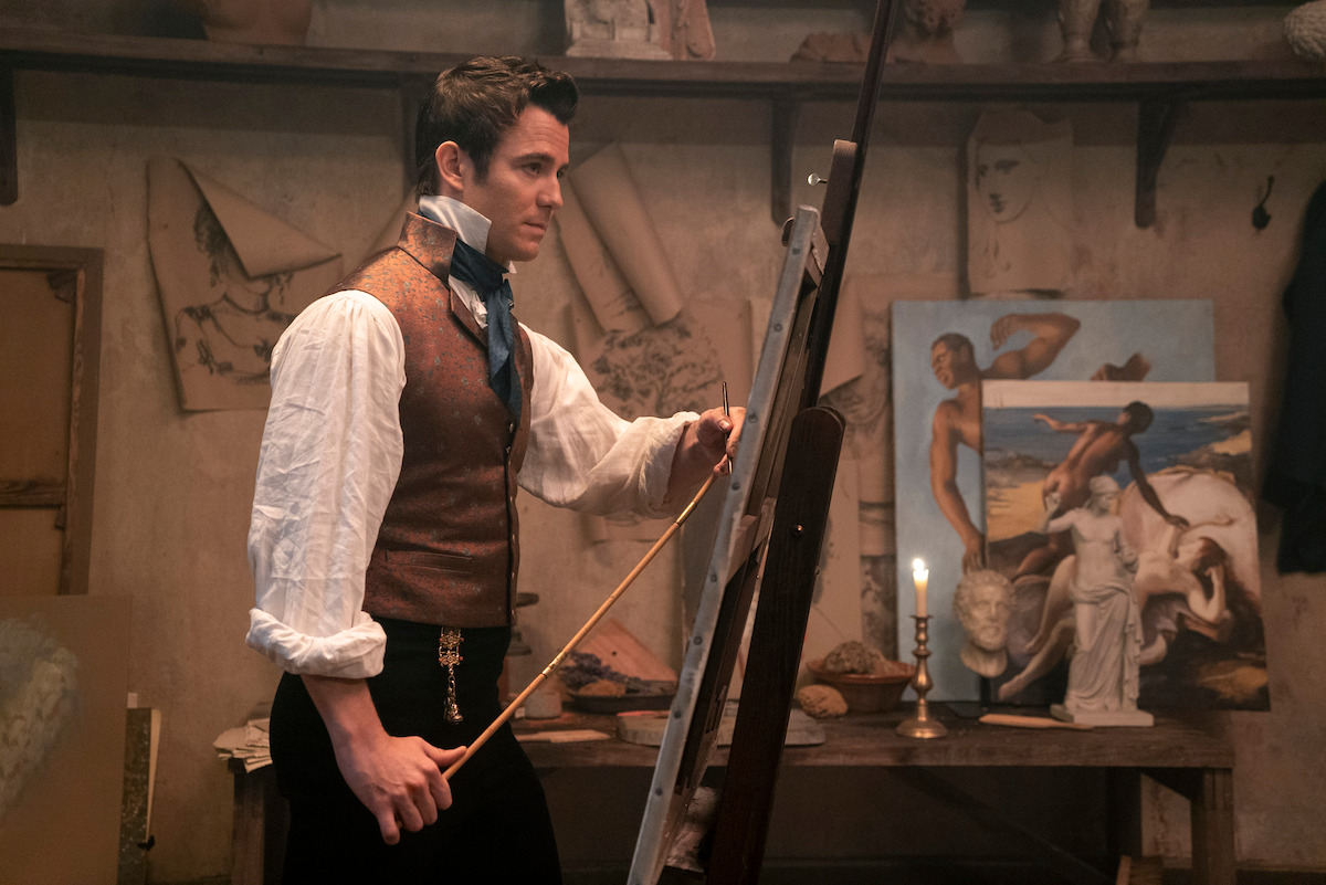A man in period clothing stands painting at an easel in a vintage art studio filled with classical sculptures, sketches, and paintings, with a lit candle on a wooden table in the background.
