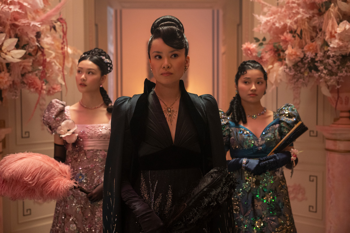 Three women in elaborate, elegant gowns stand in a richly decorated hallway with ornate floral arrangements and soft lighting, suggesting a formal or historical setting.