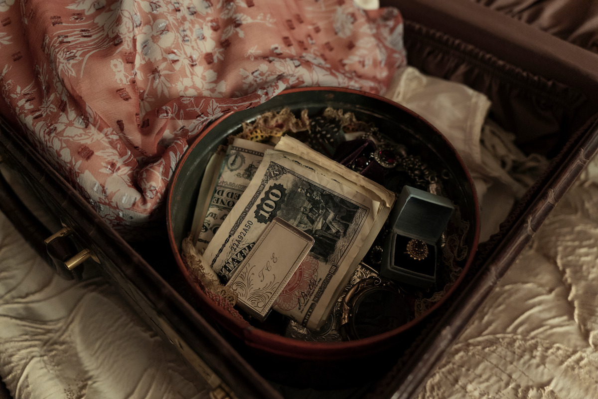 An open suitcase on a bed contains a round tin filled with vintage currency, jewelry, rings, and a pink patterned fabric, suggesting hidden valuables in a cozy, private setting.