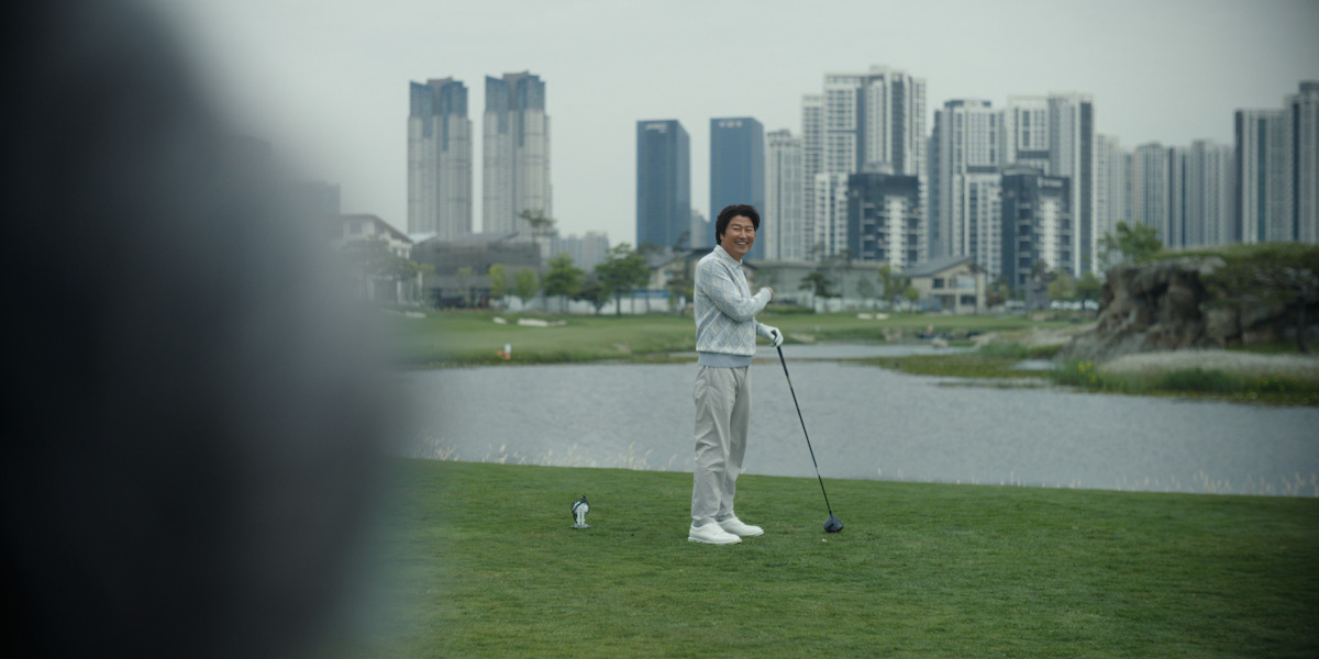 Man holding golf club on green golf course near water, with modern city skyscrapers and residential buildings in background on a cloudy day.