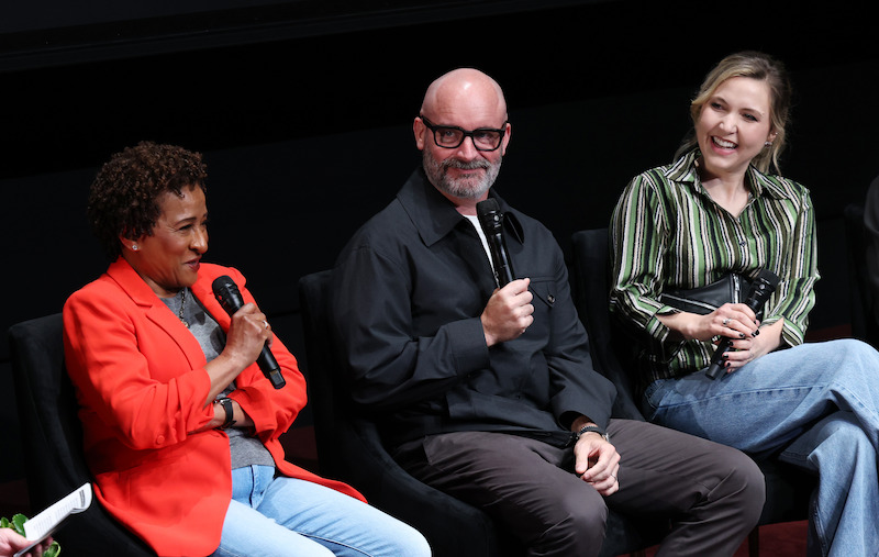Three people sit on stage in a panel discussion, each holding a microphone. They are smiling and appear engaged with the audience. The setting is indoors, with a black background and chairs in a row.