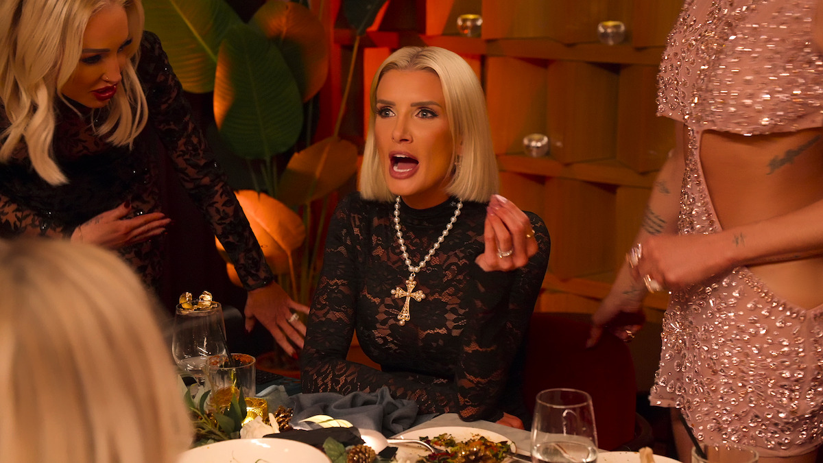 A woman in a black lace dress sits at an elegant dinner table, gesturing passionately while talking. Two other women stand nearby. The setting is warm and stylish, with dramatic lighting and a lively, intense mood.