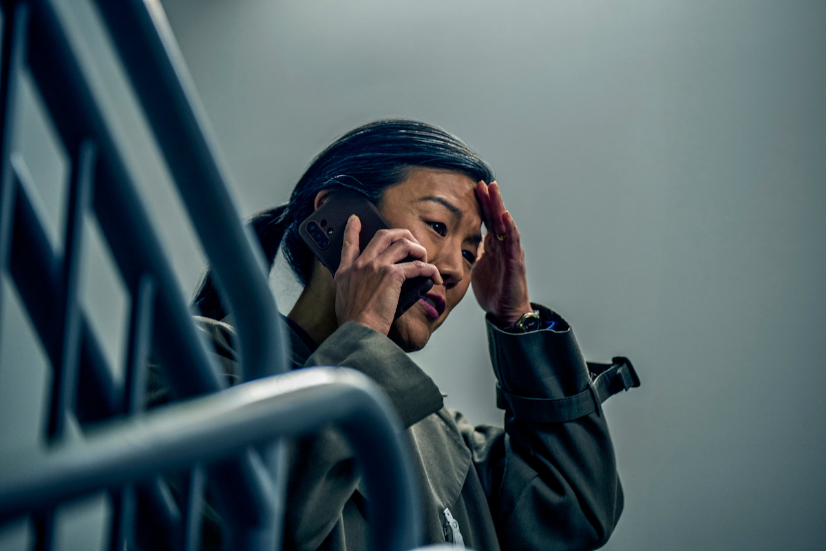 Woman on a staircase talking on the phone, looking worried and stressed, with her hand on her forehead in a dimly lit indoor environment, suggesting tension or concern.