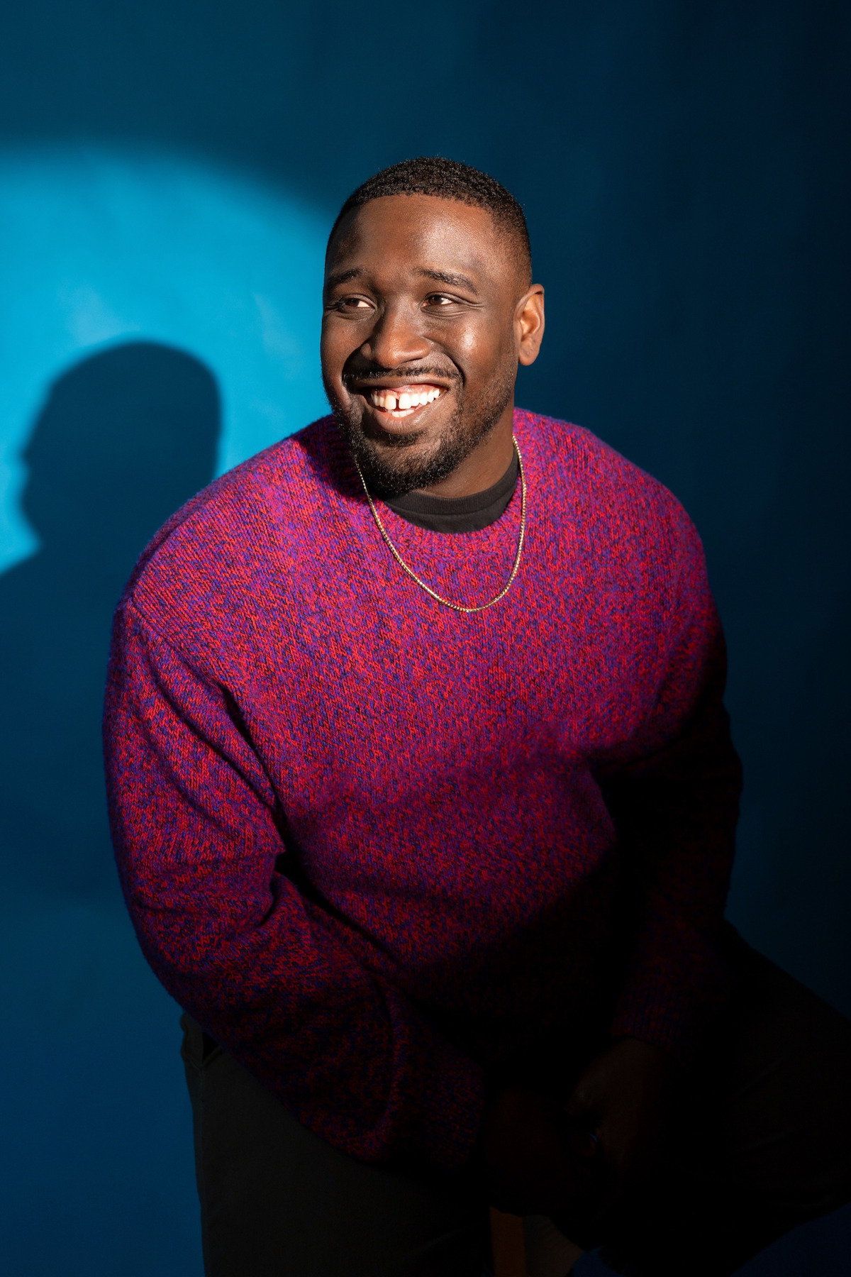 Smiling man in a red and purple sweater sits in dramatic lighting against a blue background, casting a shadow. The setting appears to be a studio environment.